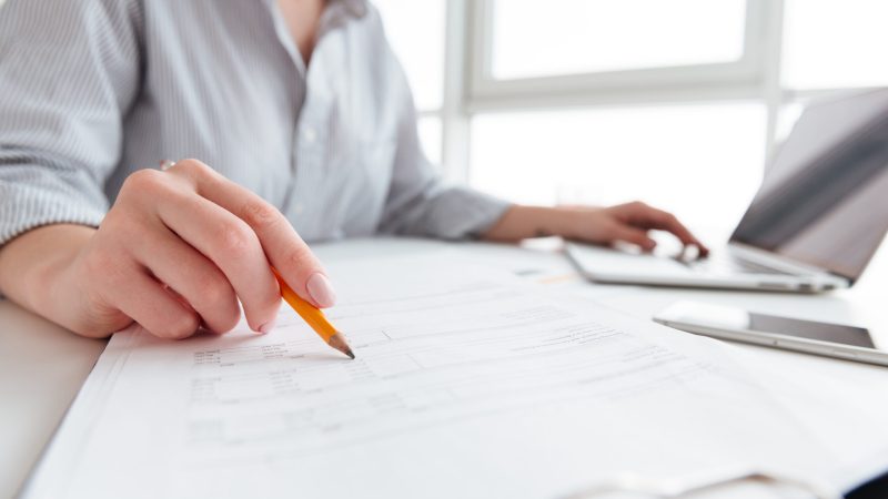 Close up portrait of a woman holding pencil over paper documents while sitting and working with laptop computer at home